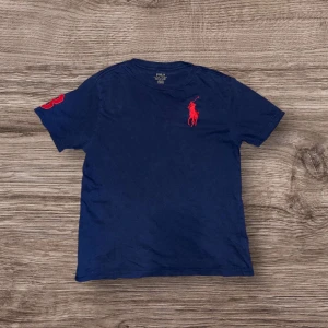 Blå t-shirt från Ralph Lauren - Säljer en snygg blå t-shirt från Ralph Lauren med röd logga på bröstet och en liten röd detalj på ärmen. Perfekt för en stilren look. Passar bra till både jeans och shorts.