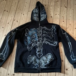 Full Zip Hoodie Travis Scott Cactus Jack For Fragment Skeleton Grafitti - Svart Travis Scott hoodie (Cactus Jack) med ett  skelettmönster i grått och blått. Tröjan har dragkedja och en stor huva som går att dra hela vägen upp, jätte cool och i bra skick. Tillverkad i 100% bomull 🥊Kan bäras av både killar och tjejer.
