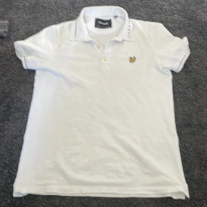 Vit pikétröja från Lyle & Scott - Säljer en klassisk vit pikétröja från Lyle & Scott med korta ärmar och en broderad logotyp på bröstet. Tröjan har en krage med knappar och är perfekt för en stilren look. Passar bra till både jeans och chinos.