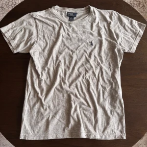 Grå Ralph Lauren t-shirt - En grå Ralph Lauren t-shirt med klassisk mörkblå logga. Den är i storlek small och i mycket bra skick. Det är en perfekt t-shirt nu när sommaren bara är runt hörnet och den passar till nästan allt och alla.