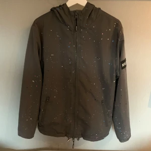 Grå jacka med färgstänk från NVLTY - Snygg grå jacka från NVLTY med unika färgstänk i olika färger. Jackan har en dragkedja framtill och en huva för extra skydd. Perfekt för att ge din outfit en cool touch.