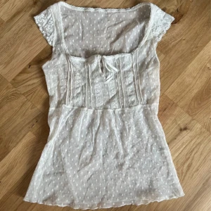 Coquette spetstopp - Charmig vit blus med spetsdetaljer och prickigt mönster. Blusen har korta ärmar och en söt knytning framtill. Perfekt för en somrig look!