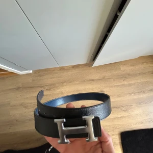 H bälte - Snyggt svart bälte med ett stort, silverfärgat H-format spänne. Strl 110. killen jag köpte av sa att det var äkta ! Bältet har en stilren design och är perfekt för att ge en extra touch till din outfit.