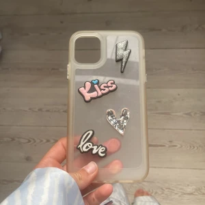 Genomskinligt mobilskal med dekorationer - Säljer ett genomskinligt mobilskal med coola 3D-dekorationer: blixt, glittrigt hjärta, texten 'Kiss' i rosa och 'love' i vitt. Skalet har en mjuk beige ton och passar perfekt om du vill ha en personlig vibe på mobilen.