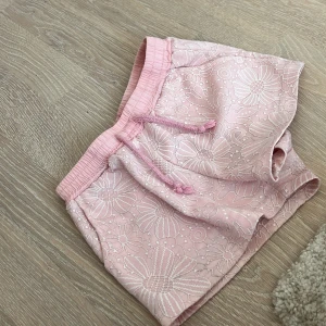 Blommiga rosa shorts med snörning - Supersöta ljusrosa shorts i storlek 134/140. Med blommor och elastisk midja med snörning. Tillverkade i mjuk polyesterblandning med lite stretch för extra komfort. Perfekta för varma sommardagar när du vill ha en chill och söt look.