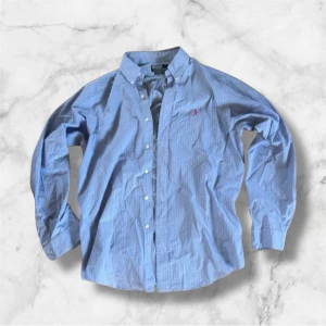 Blårutig skjorta Polo Ralph Lauren L - Klassisk blårutig skjorta från Polo Ralph Lauren i custom fit. Skjortan har button-down krage, långa ärmar och knappar framtill. Tillverkad i 100% bomull och har den ikoniska rosa loggan broderad på bröstet. Perfekt för en stilren och avslappnad look.