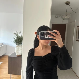 Svart glittrig kort klänning med puffärmar - Säljer en svart kort klänning med snygga puffärmar och rynkade detaljer framtill. Klänningen har en fyrkantig urringning och sitter tight över kroppen för en riktigt snygg siluett. Perfekt för dig som vill sticka ut med en stilren look. 