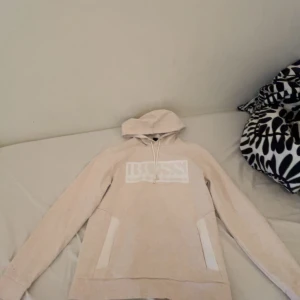 Beige hoodie från BOSS med logga - Snygg beige hoodie från BOSS med stor vit logga på bröstet. Tröjan har huva med snörning och praktiska fickor på sidorna. Perfekt för dig som gillar en clean och stilren look. Mjuk och skön bomullskänsla.