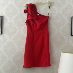 Röd oneshoulder klänning från Divided - Snygg röd klänning från Divided i oneshoulder-modell med en stor rosett på axeln. Klänningen har en figurnära passform och är tillverkad i ett mjukt syntetmaterial. Perfekt för dig som vill sticka ut med en cool och trendig look.