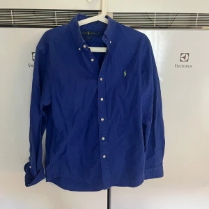 Blå skjorta från Ralph Lauren - Klassisk blå skjorta från Ralph Lauren med button-down krage och grönt broderat logotyp på bröstet. Skjortan har långa ärmar, vita knappar och är tillverkad i mjuk bomull. Perfekt för en stilren och avslappnad look.