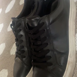 Svarta sneakers i skinn - Stilrena svarta sneakers från vagabond med ovandel i skinn och svarta snören. Skorna har en vit, platt sula som ger en klassisk look. Perfekta för dig som gillar en enkel och clean stil. Rund tå och diskret design som passar till det mesta.