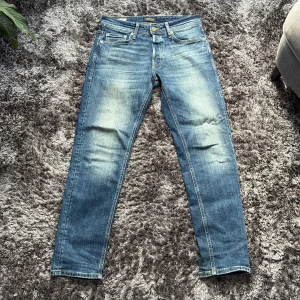 Blå Jack and Jones Jeans - Snygga blå jeans från Jack and Jones modellen tapered. Jeansen har klassisk femficksdesign, ljusa slitningar framtill och raka ben med avsmalnande passform. Tillverkade i mjukt denimtyg med gul kontrastsöm för en cool detalj.