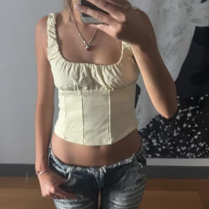 Ljusgul korsett-topp med volang - Supersöt ljusgul korsett-topp med volangkantade axelband och fyrkantig urringning. Toppen är croppad och har en figurnära passform som framhäver midjan. Perfekt att styla med jeans eller kjol för en trendig look.