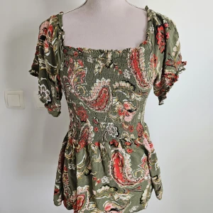 Grön blommig topp Dorothy Perkins - Supersöt topp från Dorothy Perkins i grön bas med blommigt mönster i rött, rosa och vitt. Smockad överdel, fyrkantig ringning och korta puffärmar. Tillverkad i mjuk viskos som känns lätt och skön på sommaren.