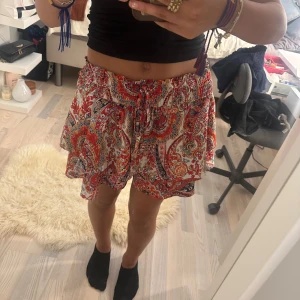 Färgglada mönstrade volang kjol - Superfina shorts med volanger och paisleymönster i rött, orange, blått och vitt. Högmidjade med resår och snörning i midjan för skön passform. Lätta och luftiga, perfekta för varma dagar. Ger en riktigt boho och chill vibe till din outfit.