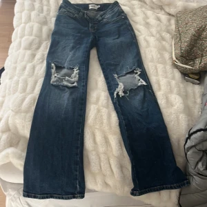 Blå jeans - Säljer ett par blå jeans med bootcut-modell och stora slitningar över knäna. Jeansen har klassisk femficksdesign, normal passform och är tillverkade i jeansmaterial. Perfekta för dig som gillar en avslappnad och edgy stil.