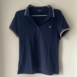 Mörkblå Fred Perry piké t-shirt - Snygg mörkblå piké från Fred Perry med klassisk krage och vita ränder på ärmslut och krage. T-shirten har korta ärmar och den ikoniska Fred Perry-loggan broderad på bröstet. Tillverkad i mjuk bomull för en skön känsla.
