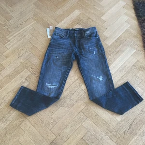 Jack & Jones blå tapered jeans W29 L32 - Snygga svarta jeans från Jack & Jones med slitna detaljer och tvättad look. Modellen är tapered med normal passform och klassisk femficksdesign. Jeansen är tillverkade i bomull och har en mörkblå färgton med coola slitningar på benen. Helt oanvända i nytt skick