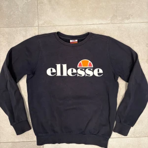 Svart sweatshirt från Ellesse - Svart sweatshirt från Ellesse med stort vitt tryck och logga i orange och rött på bröstet. Tröjan har rund hals, långa ärmar och ribbade muddar vid ärmslut och nederkant. Passar som XS