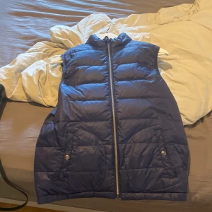 Blå dunväst från Ralph Lauren - Snygg marinblå dunväst från Ralph Lauren med klassisk broderad logga på bröstet. Västen har dragkedja framtill, två fickor med knappstängning och quiltad design. Perfekt att slänga över en hoodie för en chill look.