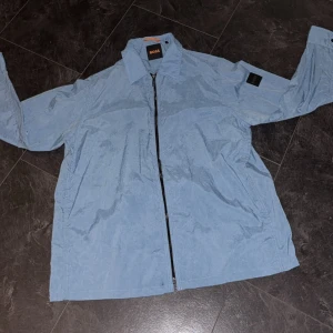 Windbreaker Hugo boss - Snygg blå vindjacka från BOSS med klassisk krage och hel dragkedja framtill. Jackan har en stilren look med diskret BOSS-logga på ärmen och är tillverkad i lätt syntetmaterial. Perfekt för dig som vill ha en clean och modern stil.