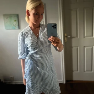 Blåvit omlottklänning med knyt - Säljer en fräsch kort omlottklänning med blå och vita vertikala ränder. Klänningen har trekvartsärm, v-ringad hals och knyt i midjan för en snygg passform. Materialet är lätt och känns som bomull, perfekt för varma dagar. Supersöt och enkel att styla!