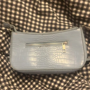 Ljusblå axelväska i crocopräglat syntet - Säljer en ljusblå handväska för 80kr. Har använt den några gånger men sållar så kommer jag göra dne fin innan den ska levereras