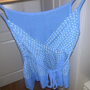 Blå prickig topp med knyt - Säljer en söt blå topp med vita prickar och tunna axelband. Toppen har omlott-design framtill och ett knyt i midjan för extra passform. Perfekt för varma dagar och har en lätt, flowy känsla. Materialet känns mjukt och bekvämt mot huden. Man kan knyta banden fram och bak  