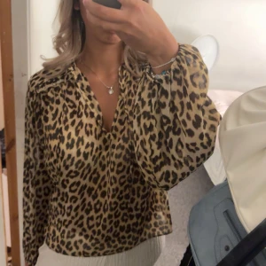 Leopardmönstrad blus med v-ringning - Säljer en snygg blus med leopardmönster i beige och svart. Blusen har v-ringad hals och långa, luftiga ärmar med resår vid handleden finns två band så man kan knyta om man inte vill ha de så urringat . Perfekt för dig som vill sticka ut med ett djuriskt print och skön passform.