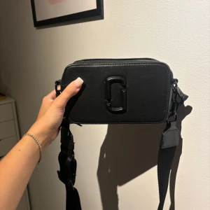 Svart axelväska från Marc Jacobs - Säljer en svart axelväska från Marc Jacobs med stilren design och bred justerbar rem. Väskan har två dragkedjor upptill, präglad logga på sidan och ett snyggt emblem framtill. Materialet är skinn med en lätt textur och väskan är rektangulär i formen.