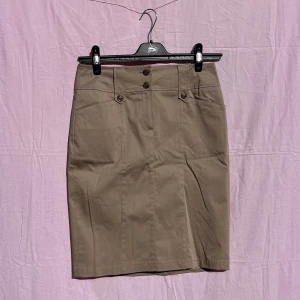 Beige pennkjol från Taifun - Snygg beige pennkjol från Taifun med hög midja och dekorativa knappar framtill. Kjolen har fickor fram och bälteshällor, perfekt för en clean och stilren look. Materialet är mjukt och känns bekvämt mot huden.