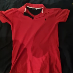 Röd Polo Ralph Lauren pikétröja äkta - Snygg röd pikétröja från Polo Ralph Lauren med klassisk krage och svart broderad logga på bröstet. Tröjan är kortärmad och tillverkad i mjuk bomull, perfekt för en clean och sportig look.