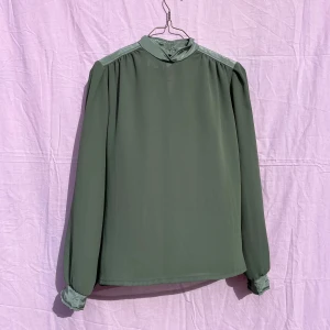 Grön blus från Mac Scott, stl 36 - Snygg ljusgrön blus från Mac Scott med hög krage och knäppning i nacken. Blusen har långa ärmar med satinliknande detaljer på axlar och ärmslut. Tillverkad i 100% polyester och har en stilren, rak passform. Perfekt för dig som gillar en clean och elegant look.