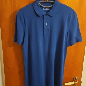 Blå pikétröja från Dressmann L - Snygg blå pikétröja från Dressmann i storlek L. Klassisk modell med krage och två knappar framtill. Tillverkad i mjuk bomull som känns skön mot huden. Perfekt för en clean och stilren look.