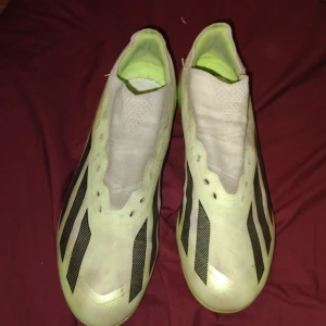 Adidas fotbollsskor i ljusgrönt - Snygga fotbollsskor från Adidas i ljusgrönt och vitt med svarta ränder på sidorna. Skorna har en strömlinjeformad design utan snörning och är tillverkade i syntetmaterial för bästa känsla på planen. Perfekta för dig som vill sticka ut på matchen.