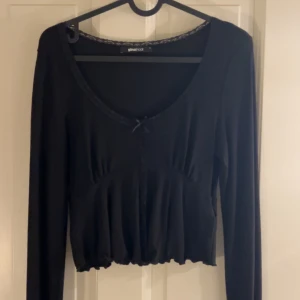 Svart långärmad topp från Gina Tricot - Supersöt svart långärmad topp från Gina Tricot med croppad passform och liten rosett framtill. Djup rundringning och lätt rynkad nedtill för en feminin vibe. Perfekt att styla med jeans eller kjol för en trendig look.