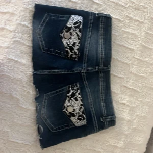 Mörkblå jeanskjol med snake-detaljer - Snygg mörkblå jeanskjol med råa kanter och kontrastsömmar. Baksidan har coola fickor med svartvita ormmönstrade detaljer och silvriga knappar. Perfekta för dig som vill sticka ut lite extra i sommar! Klassisk femficksmodell och dragkedja framtill.