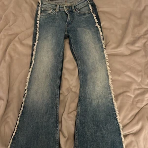 Blå bootcut jeans med fransade kanter - Säljer ett par blå bootcut jeans med ljus tvätt och råa, fransade kanter längs sidorna och midjan. Jeansen har klassisk femficksdesign och bälteshällor. Perfekta för dig som vill sticka ut med en edgy vibe.