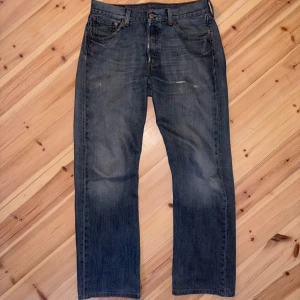 Levi's 501 raka jeans blå denim - Klassiska Levi's 501 jeans i blå denim med raka ben och normal passform. Jeansen har fem fickor, bälteshällor och dragkedjegylf. Snyggt slitna detaljer och ikonisk läderpatch bak i midjan. Perfekta för en avslappnad streetstil.