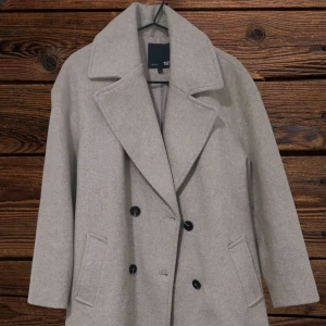 Beige kappa från 157 med stora slag - Stilren beige kappa från 157 med dubbelknäppning och stora slag. Jackan har två fickor framtill och är helfodrad. Perfekt för dig som gillar en klassisk och clean look under höst och vinter. Den är lite nopprig men har försökt tagit bort det mesta.