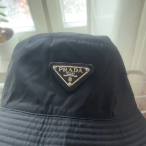 Svart bucket hat från Prada - Snygg svart bucket hat från Prada med ikonisk triangellogo framtill i metall och läder. Hatten är tillverkad i ett glansigt syntetmaterial som ger en exklusiv känsla. Perfekt accessoar för att lyfta din outfit med en lyxig touch.