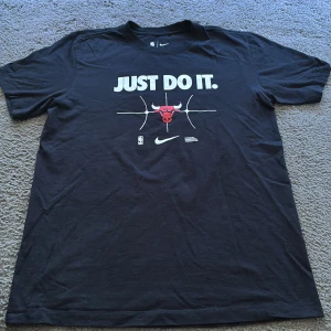 Svart Nike x NBA Bulls t-shirt - Svart t-shirt från Nike med Chicago Bulls-logga och stor vit 'JUST DO IT.' text på bröstet. Rund hals, korta ärmar och basketbollgrafik. Materialet är mjuk bomull, perfekt för dig som gillar basket och streetwear.