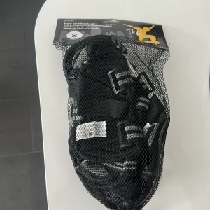 Svarta skatehandskar SK8 strl S - Svarta handskar från SK8, perfekta för skate eller inlines. De har justerbara remmar med spännen och är tillverkade i slitstarkt syntetmaterial. Säljs i ett praktiskt nätfodral som gör dem enkla att ta med till skateparken.