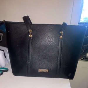 Svart axelväska från Anna Field - Stilren svart axelväska från Anna Field med guldiga metalldetaljer och dubbla handtag. Väskan är tillverkad i skinnimitation och har en klassisk, rymlig form som passar perfekt för att bära med sig allt man behöver. Snygg och enkel att matcha till olika outfits. Den är perfekt som skolväska och praktisk att ta med sig överallt. 