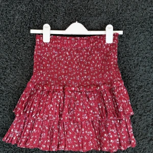 Röd blommig volangkjol Isabel Marant - Supersöt röd kortkjol från Isabel Marant Étoile med små blommor i vitt och lila. Kjolen har smockad midja och flera volanger som ger en flowy känsla. Tillverkad i mjuk bomull, perfekt för vår och sommar. Modellen heter Naomi.