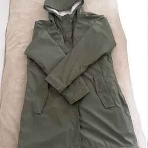 Olivgrön regnjacka med teddyfoder  - Olivgrön regnjacka med vit teddyfoder på insidan. Jackan har huva, dragkedja och tryckknappar framtill samt två stora fickor. Perfekt för kalla och regniga dagar när du vill hålla dig både varm och torr.