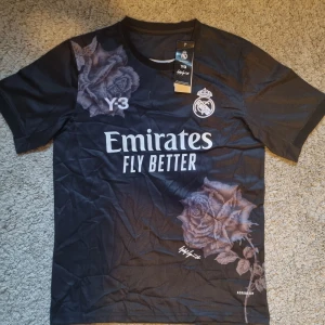 Real Madrid x Y-3 svart fotbollströja - Snygg svart Real Madrid fotbollströja från Y-3 kollektionen, designad för klubbens 120-årsjubileum. Tröjan har korta ärmar, stora blommiga rosor i grå toner och klassiska loggor framtill. Tillverkad i lätt och ventilerande material med Aeroready-teknologi.