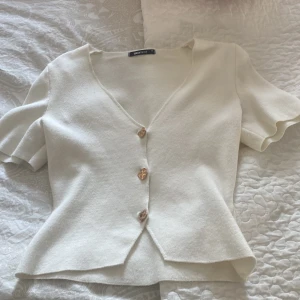 Vit v-ringad blus från Gina Tricot - Supersöt vit blus från Gina Tricot i storlek XS. Den har v-ringning, korta ärmar och tre snygga guldfärgade knappar framtill. Materialet är mjukt och känns lite stickat, perfekt för en chill och stilren look. Passformen är normal och toppen är lätt croppad.