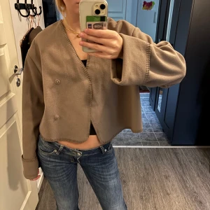 Brun/Beige cardigan jacka från Gina Tricot - Säljer en cardigan jacka från Gina Tricot med croppad passform och breda, långa ärmar. Jackan har dekorativa knappar framtill och synliga sömmar längs kanterna.