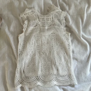 Vit spetsblus med detaljerad brodyr - Superfin vit blus i spets med massor av broderade detaljer och genombrutet mönster. Ärmlös modell med vågig kant nertill och vid axlarna. Perfekt för dig som gillar romantisk och bohemisk stil. Snygg att styla med jeans eller kjol.
